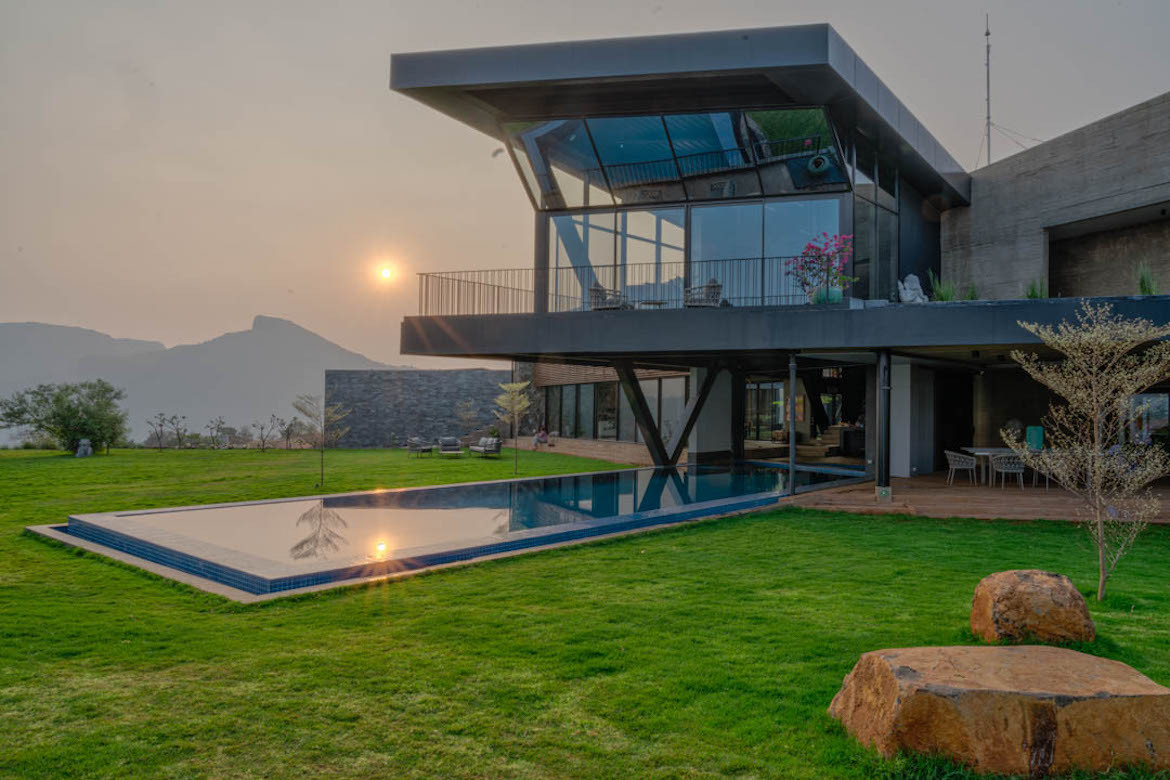 Airavat, a cantilevered modern home extends above a pool and green grass. A fog-covered sun hovers above a mountain the background.