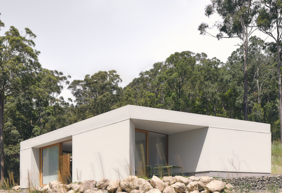 A single-storey oblique white house sits under tall woodland.