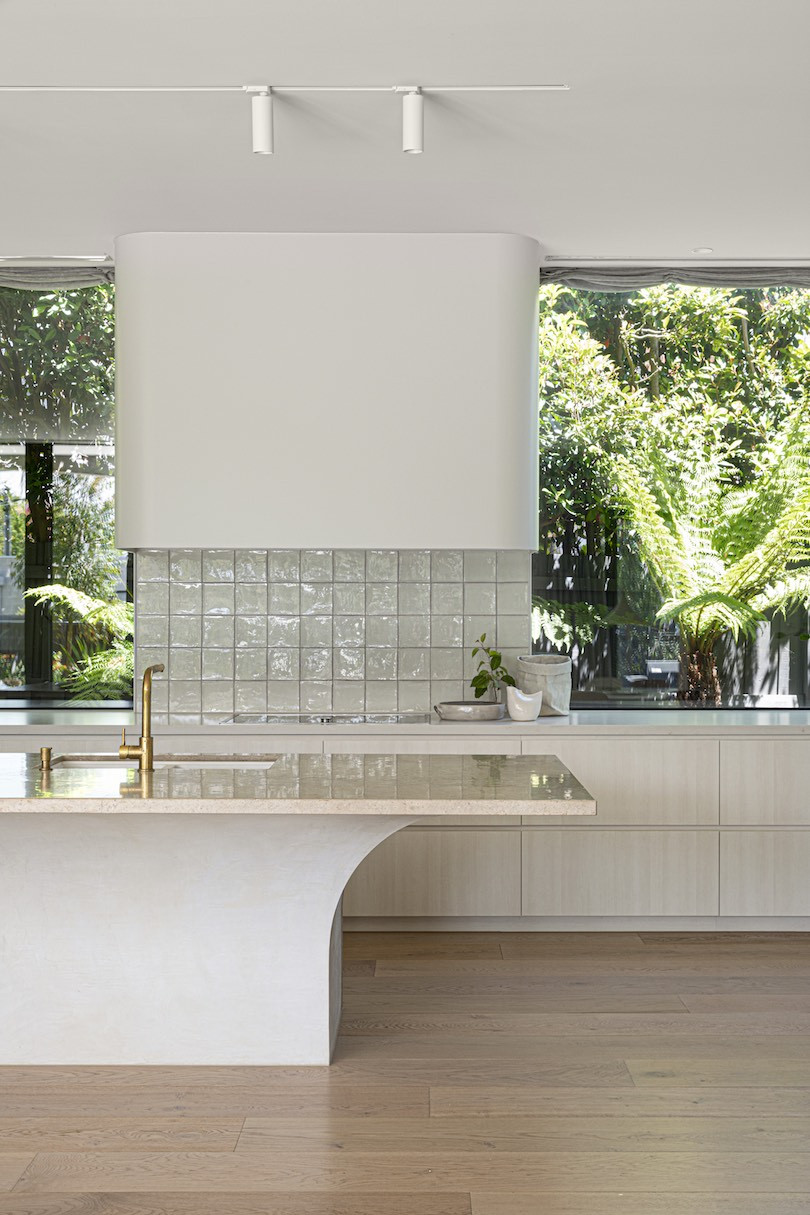 A white kitchen with green through the windows and an island bench with a curved base in brush house.