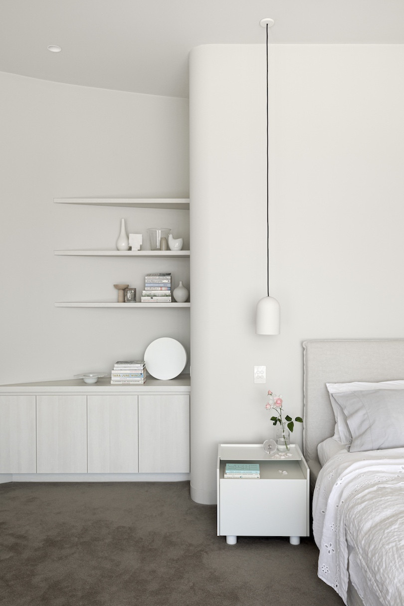 A lamp dangles from the ceiling next to a partially visible bed, a curved wall and custom white shelves in Brush House.