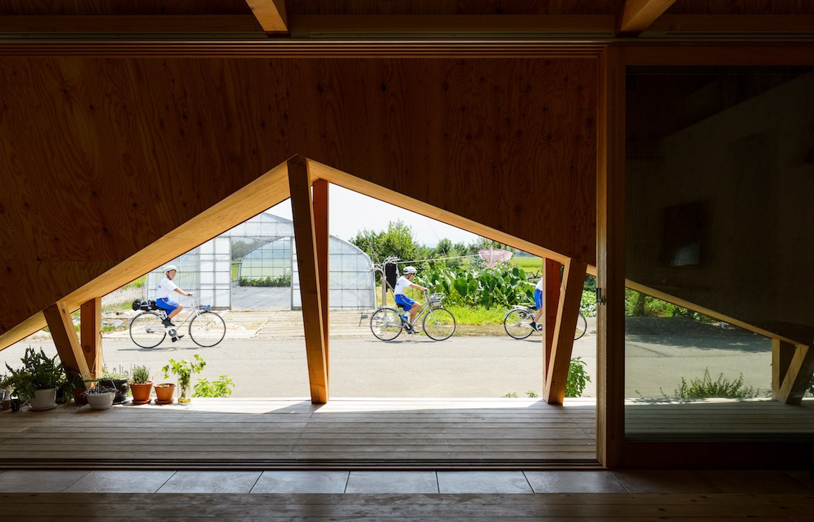 Looking out under the triangular eaves of Hara House towards a greenhouse as three kids in uniform ride past.