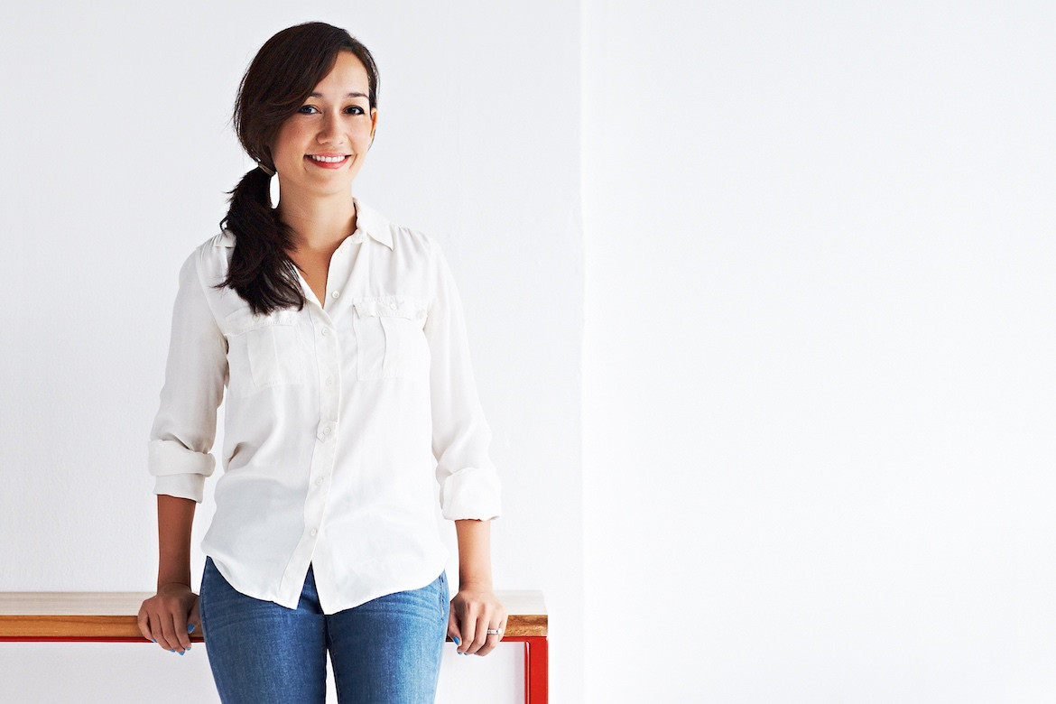 Reddie founder Caroline Olah leans on a red table in front of a white wall while wearing a white shirt and jeans.