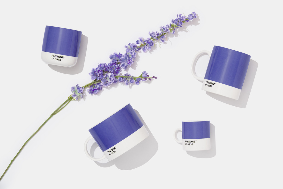 Four Pantone mugs in the colour Very Peri, a periwinkle blue, flat laid beside a sprig of lavendar.