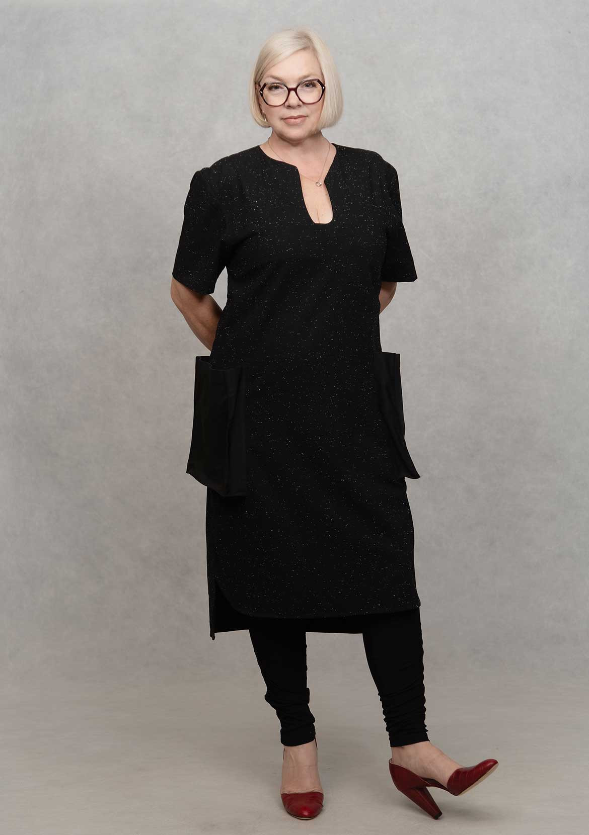 Christine McKanna-Farr of designmilkstudio stands, wearing a black dress and red heels in front of a grey background.