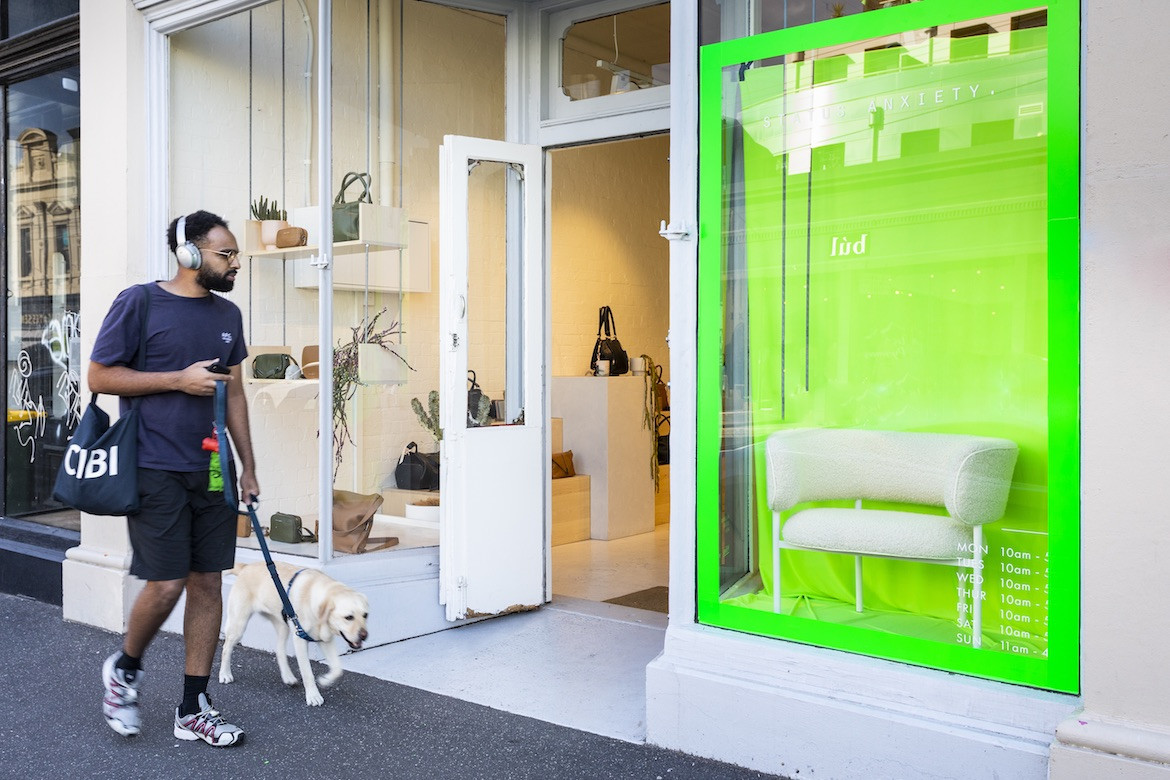 A person in a navy blue shirt and black pants walks a dog outside a shop that has a bright green display and a white chair in its window.