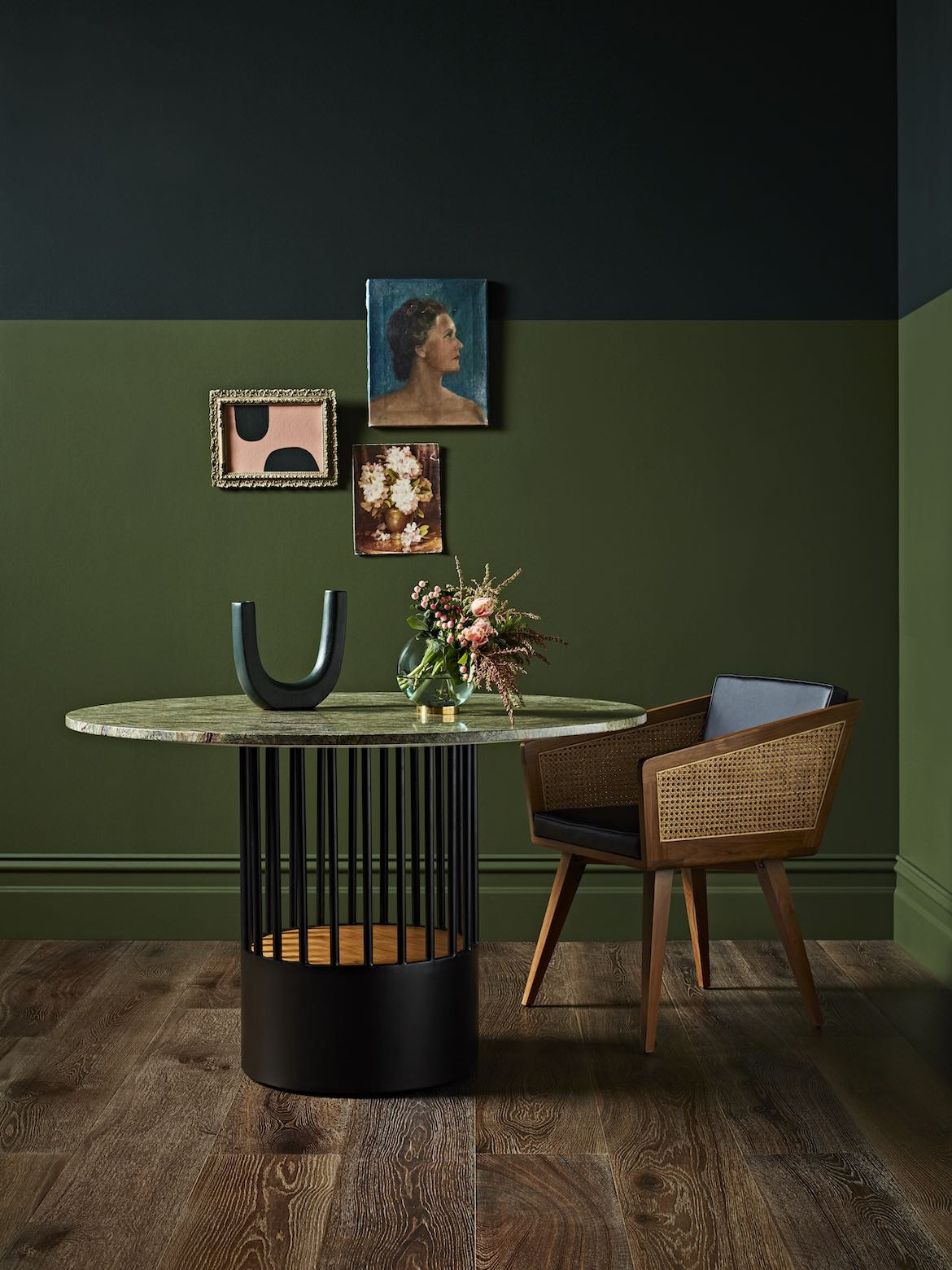 An angular black and rattan Reddie chair next to a circular green table with a cylindrical base in front of a green wall.