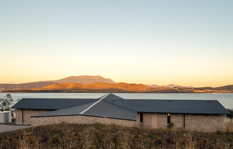 A view over the charcoal-grey roof of A woman sits on the large window frames looking out over the water in Thinking Paddock House by Open Creative Studio and out to the water.