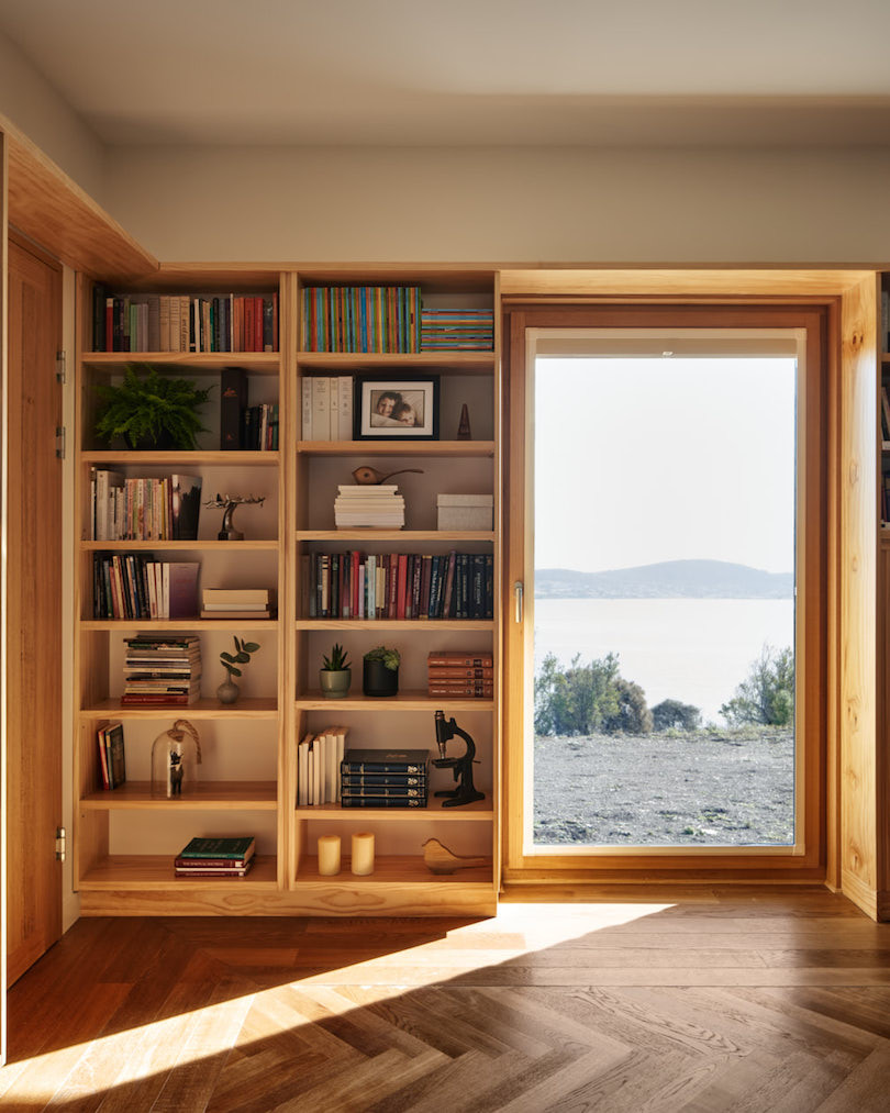 Bookshelves and a glass door with a view over the water in Four large windows with a triangle roof looking out over the bay from Thinking Paddock House by Open Creative Studio.