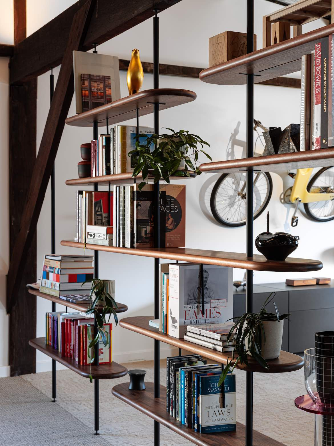 Elan Plus Morse brown timber Shelving with black beams, holding books and plans, in front of a yellow bike hanging on a wall above a couch.